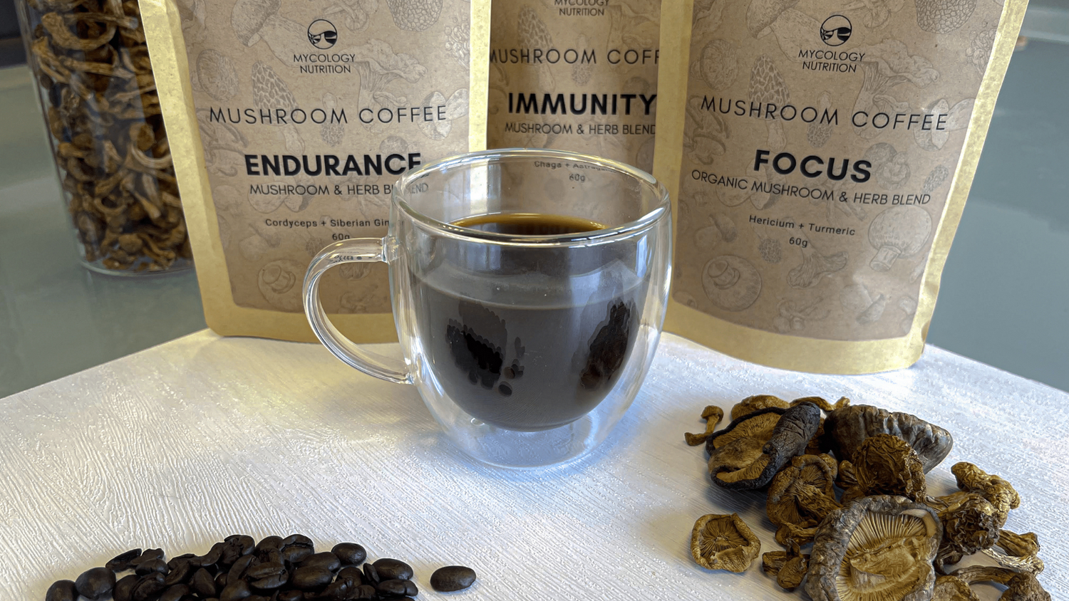 A glass cup of mushroom coffee with Mycology Nutrition blends, coffee beans and dried mushrooms.