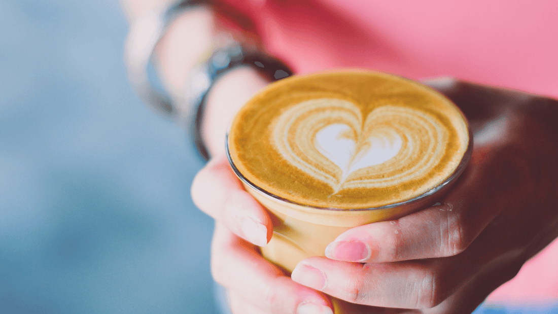 A person holding a cup of coffee with heart-shaped latte art on the surface.